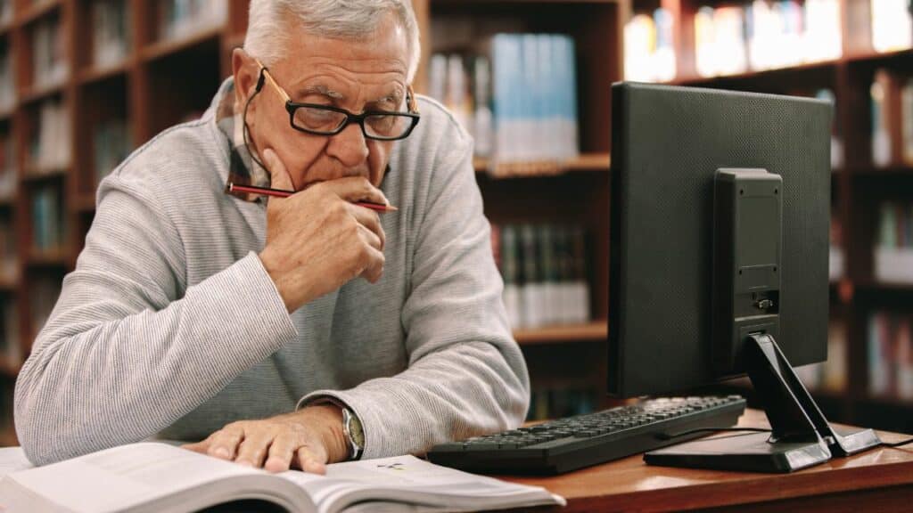 older man studying textbook at a computer