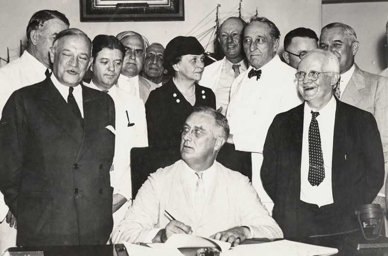 President Roosevelt signs the Social Security Act with a group of senators, representatives, and secretaries behind him.