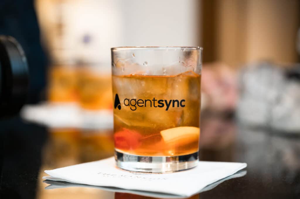 close-up cocktail glass half-full with a brown liquor and garnish and the AgentSync logo