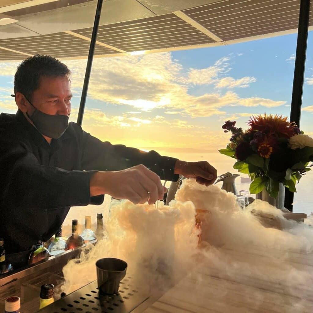 Photo of a mixologist stirring two smoky drinks at sunset in the space needle.